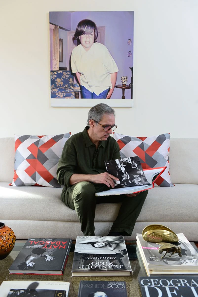 Adrián Brown sentado en el sillón con almohadones con estampas geométricas, con sus coffee table books, y arriba la pintura de Diego Gravinese. 